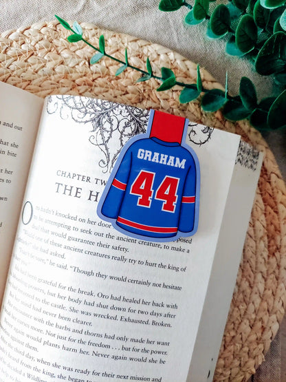 Off Campus inspired hockey jersey magnetic bookmark clipped onto an open book, showing the blue and red jersey design with "Graham 44" on the back.