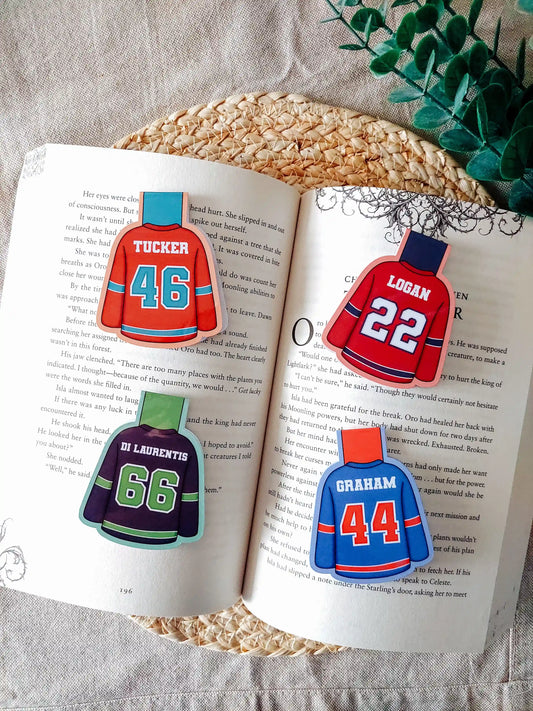 Off Campus inspired hockey jersey magnetic bookmarks displayed on an open book, featuring "Tucker 46", "Logan 22", "Di Laurentis 66" and "Graham 44" in their book cover colours. 