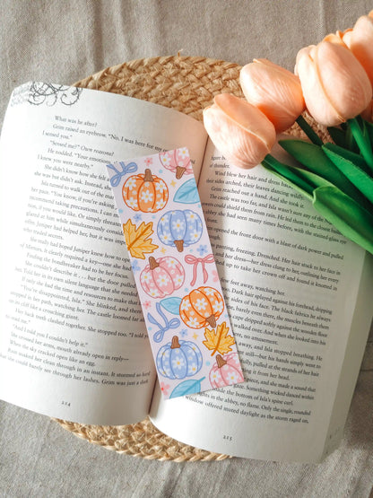 A laminated bookmark featuring illustrated pumpkins in pastel orange, pink, and blue, decorated with small white daisies, autumn maple leaves, and ribbons on a pale pink background, resting on an open book on a woven rattan mat, with peach tulips in the upper right corner.