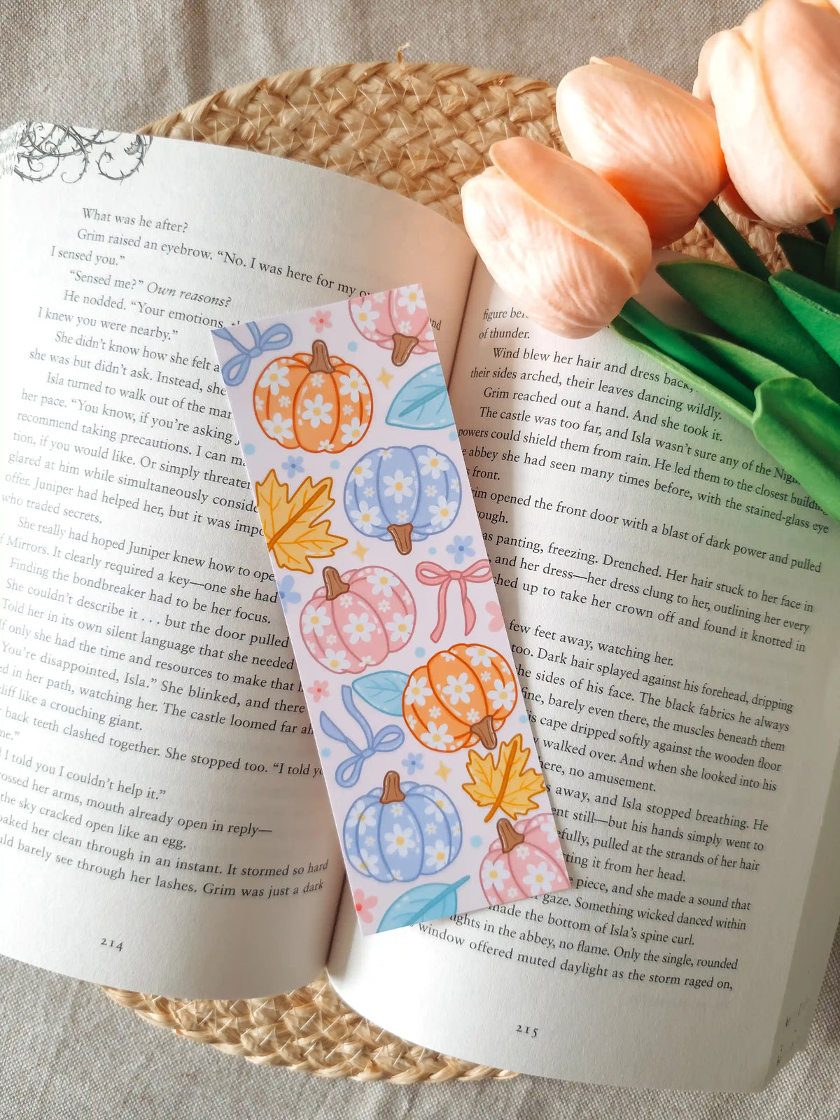 A laminated bookmark featuring illustrated pumpkins in pastel orange, pink, and blue, decorated with small white daisies, autumn maple leaves, and ribbons on a pale pink background, resting on an open book on a woven rattan mat, with peach tulips in the upper right corner.