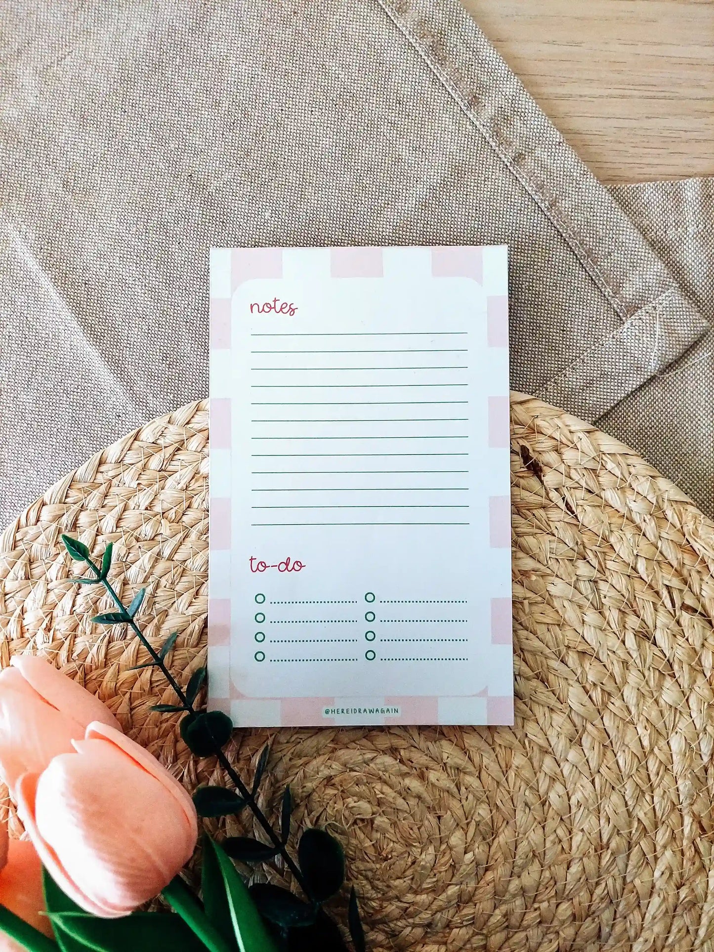Pink checkered Notes and to-do notepad featuring a lined notes section at the top and a checklist-style to-do section at the bottom, photographed on a woven surface with decorative flowers.