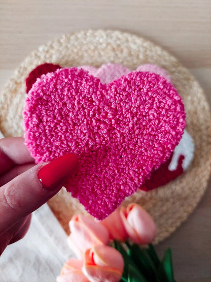 Handmade punch needle heart coaster in pink, photographed on a woven mat with decorative flowers.
