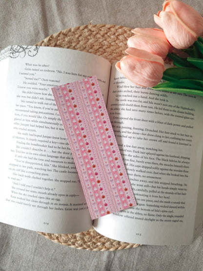 A laminated pink bookmark with a striped wavy pattern featuring illustrated cute white ghosts, small orange pumpkins, pink bows, and stars, resting on an open book on a woven rattan mat, with peach tulips visible in the upper right corner.