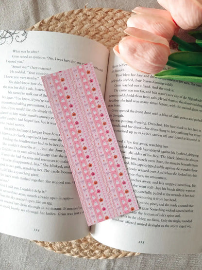 A laminated pink bookmark with a striped wavy pattern featuring illustrated cute white ghosts, small orange pumpkins, pink bows, and stars, resting on an open book on a woven rattan mat, with peach tulips visible in the upper right corner.