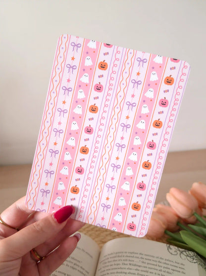 A laminated e-reader insert on a pink background with vertical stripes featuring illustrated cute white ghosts, jack-o'-lantern pumpkins in orange and pink, lavender and pink bows, candy, and stars, resting on an open book on a woven rattan mat, with peach tulips and green leaves in the upper left corner.