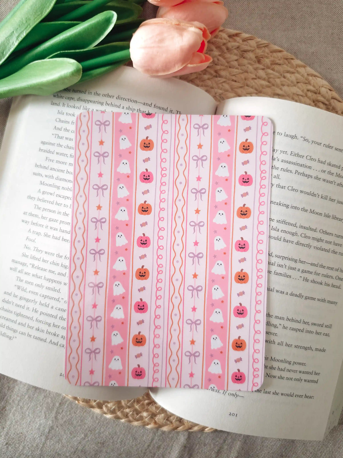 A laminated e-reader insert on a pink background with vertical stripes featuring illustrated cute white ghosts, jack-o'-lantern pumpkins in orange and pink, lavender and pink bows, candy, and stars, resting on an open book on a woven rattan mat, with peach tulips and green leaves in the upper left corner.