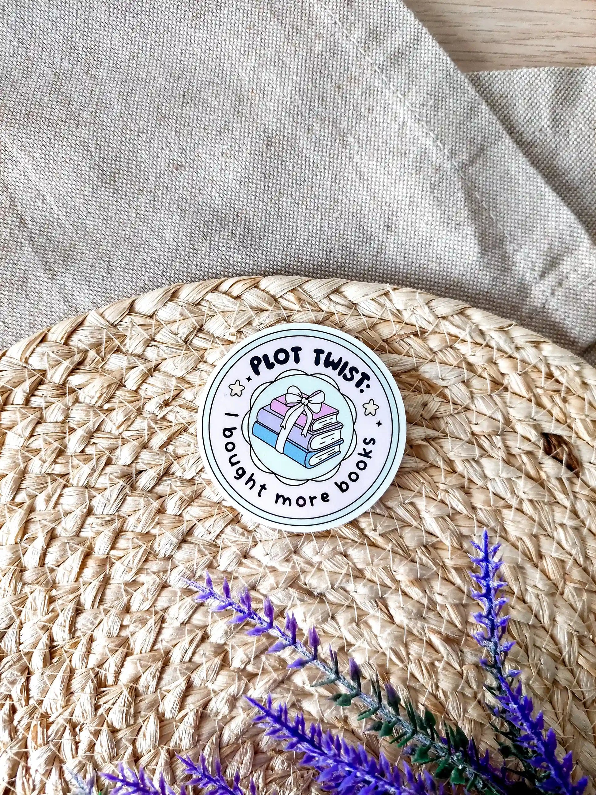 Round vinyl sticker with the text "Plot twist I bought more books" with pastel stacked books tied with a bow, photographed on neutral fabric background with woven mat and lavender.