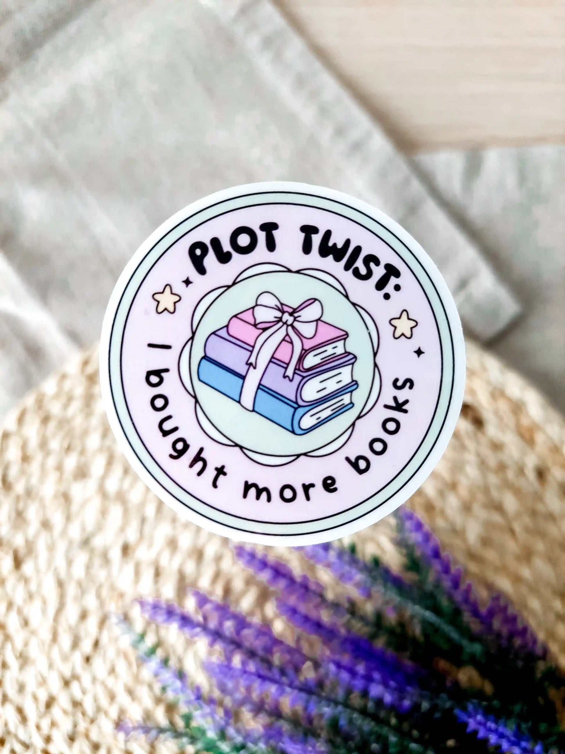 Round vinyl sticker with the text "Plot twist I bought more books" with pastel stacked books tied with a bow, photographed on neutral fabric background with woven mat and lavender.