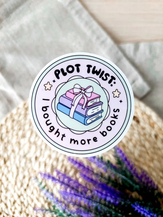 Round vinyl sticker with the text "Plot twist I bought more books" with pastel stacked books tied with a bow, photographed on neutral fabric background with woven mat and lavender.