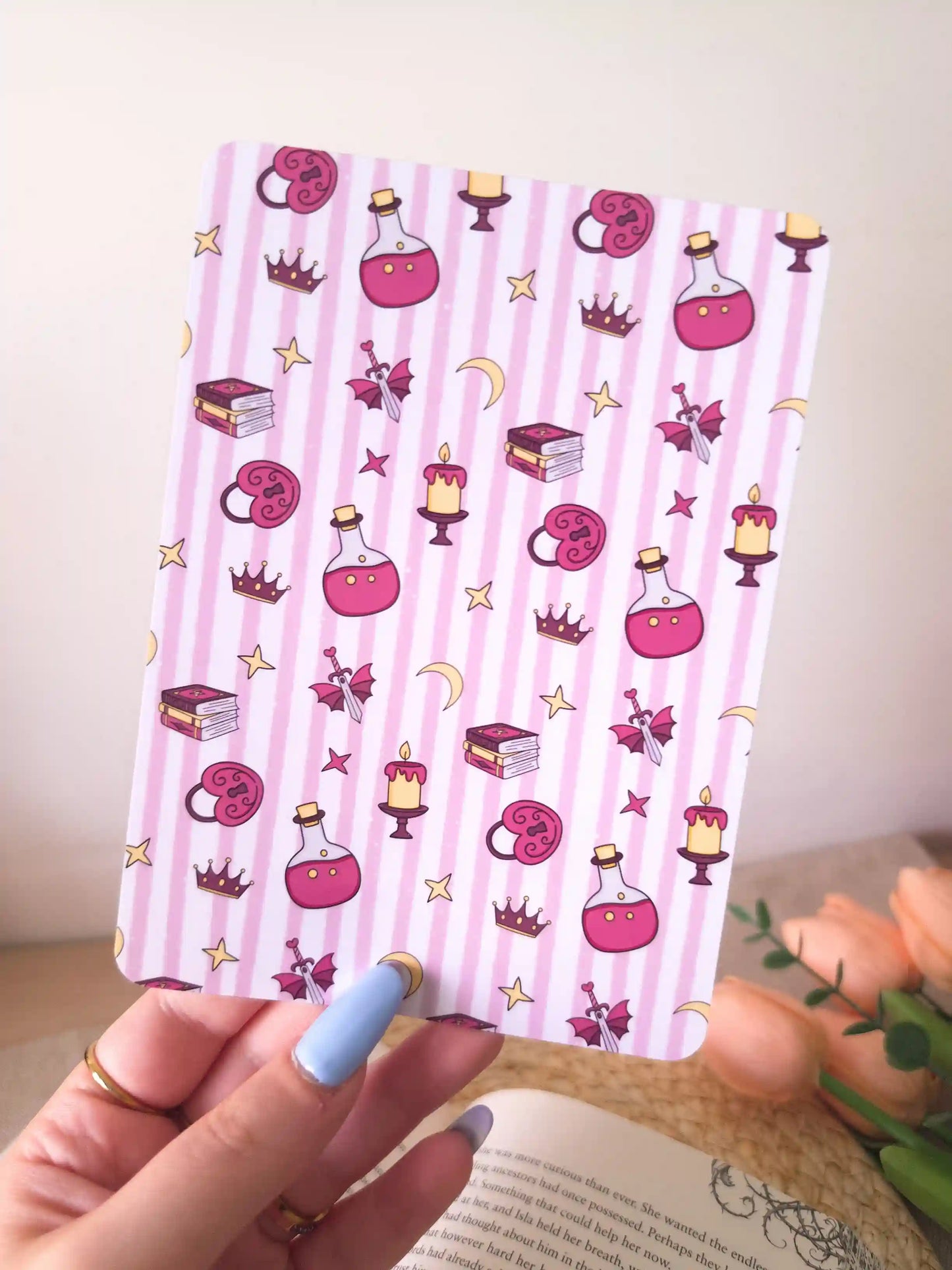 A hand holding a pink and white striped e-reader insert with a whimsical pattern of pink books, potion bottles, candles, stars, and crowns.