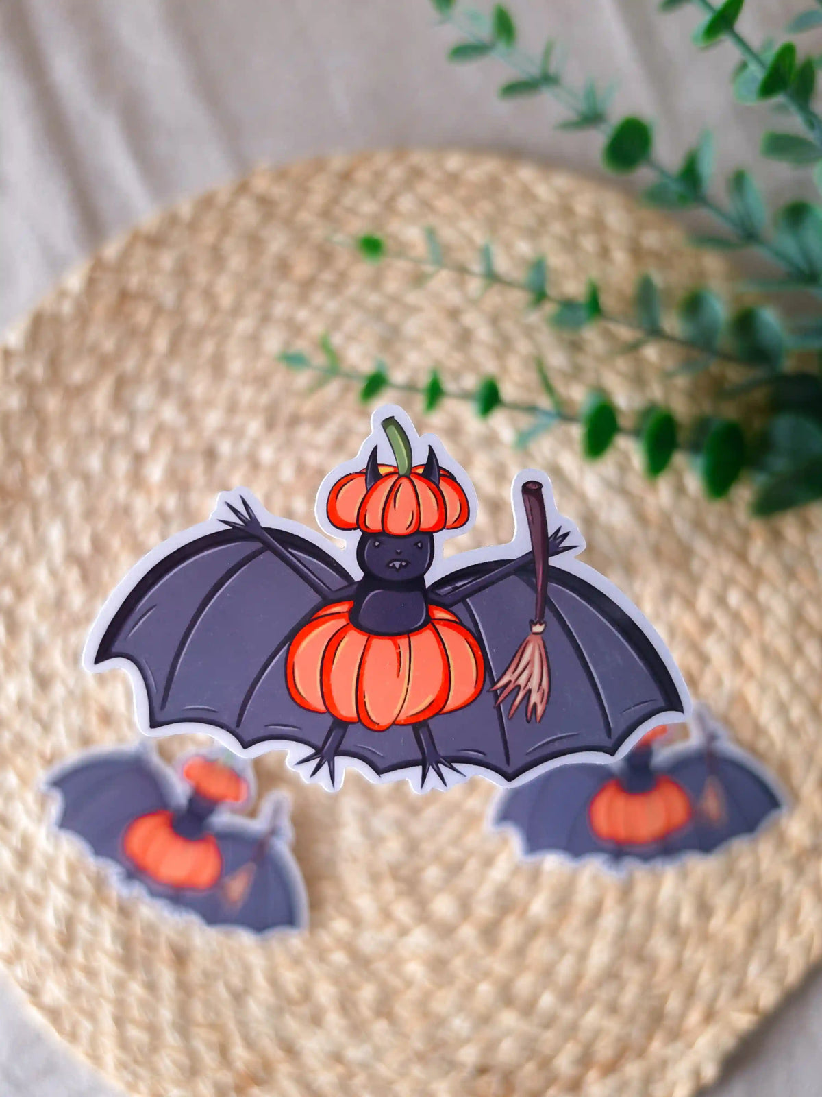A large die-cut glossy vinyl sticker of a dark purple bat wearing an orange pumpkin as a costume, with a matching pumpkin-shaped hat and a wooden broomstick in one wing, displayed alongside two smaller versions of the same sticker on a woven rattan mat, with green eucalyptus sprigs in the upper right corner.