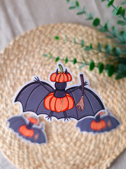 A large die-cut glossy vinyl sticker of a dark purple bat wearing an orange pumpkin as a costume, with a matching pumpkin-shaped hat and a wooden broomstick in one wing, displayed alongside two smaller versions of the same sticker on a woven rattan mat, with green eucalyptus sprigs in the upper right corner.