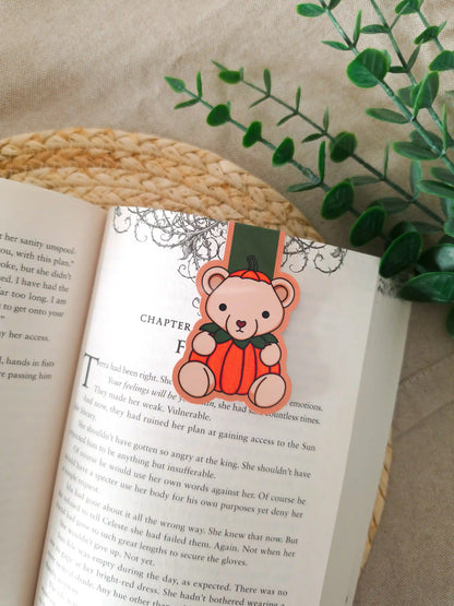 A die-cut magnetic bookmark featuring an illustrated beige teddy bear wearing a bright orange pumpkin costume with green leaves at the collar, clipped onto the top corner of an open book resting on a woven rattan mat, with green eucalyptus sprigs in the upper right corner.