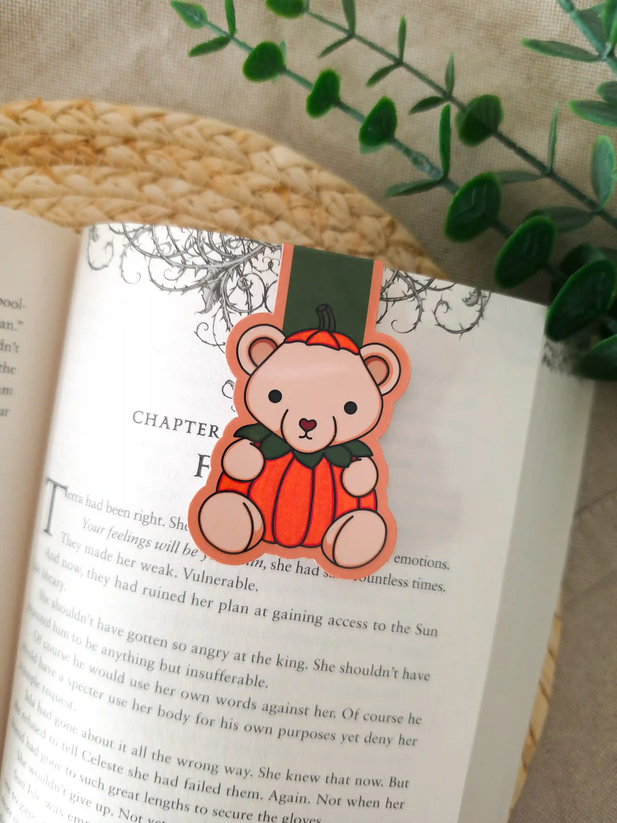 A die-cut magnetic bookmark featuring an illustrated beige teddy bear wearing a bright orange pumpkin costume with green leaves at the collar, clipped onto the top corner of an open book resting on a woven rattan mat, with green eucalyptus sprigs in the upper right corner.