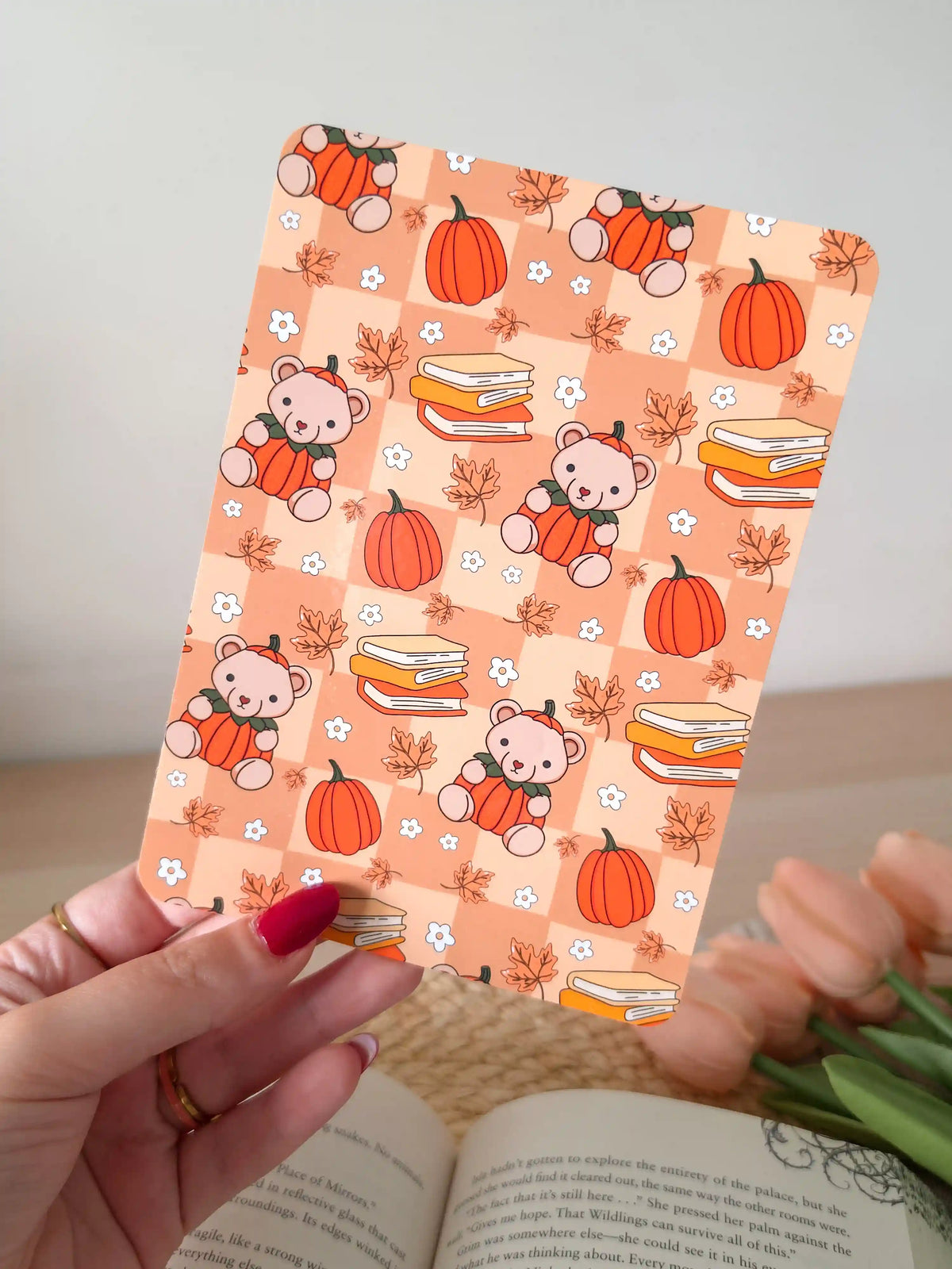 A laminated e-reader insert with a warm peach checkered background featuring illustrated teddy bears wearing pumpkin costumes, stacks of colorful books, orange pumpkins, autumn maple leaves, and small white daisies, resting on an open book on a woven rattan mat, with green eucalyptus sprigs in the upper right corner.