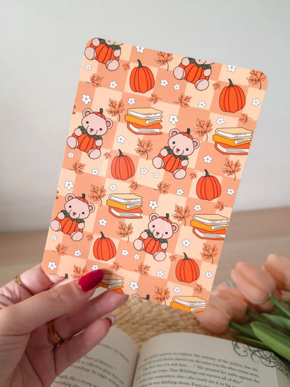 A laminated e-reader insert with a warm peach checkered background featuring illustrated teddy bears wearing pumpkin costumes, stacks of colorful books, orange pumpkins, autumn maple leaves, and small white daisies, resting on an open book on a woven rattan mat, with green eucalyptus sprigs in the upper right corner.