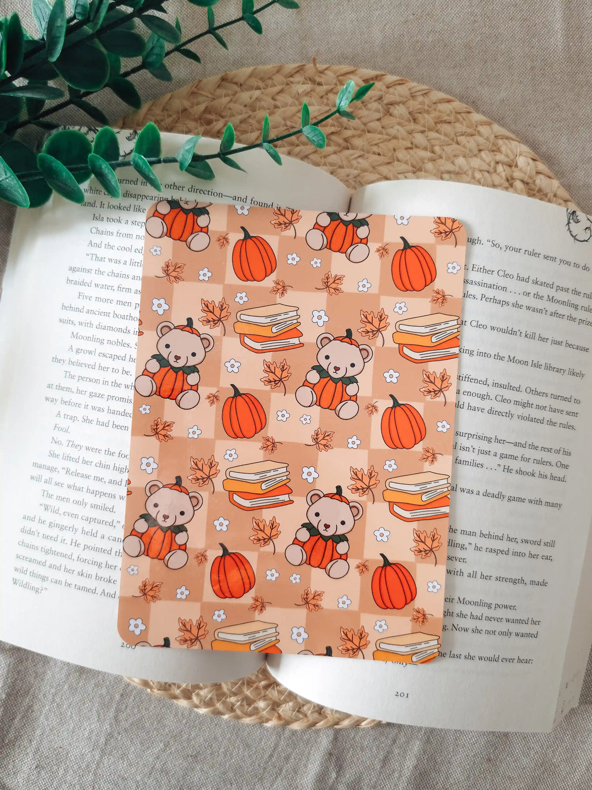 A laminated e-reader insert with a warm peach checkered background featuring illustrated teddy bears wearing pumpkin costumes, stacks of colorful books, orange pumpkins, autumn maple leaves, and small white daisies, resting on an open book on a woven rattan mat, with green eucalyptus sprigs in the upper right corner.