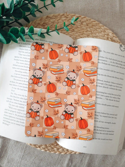 A laminated e-reader insert with a warm peach checkered background featuring illustrated teddy bears wearing pumpkin costumes, stacks of colorful books, orange pumpkins, autumn maple leaves, and small white daisies, resting on an open book on a woven rattan mat, with green eucalyptus sprigs in the upper right corner.