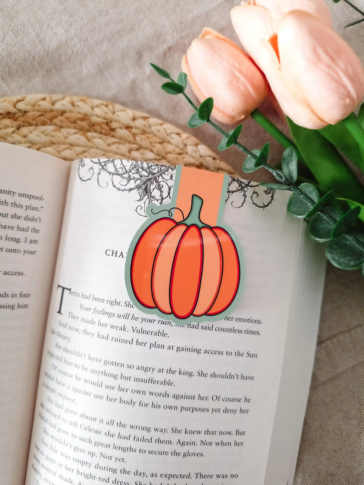 A die-cut magnetic bookmark in the shape of a round orange pumpkin with a green stem and dark outline, clipped onto the top corner of an open book resting on a woven rattan mat, with peach tulips and green eucalyptus sprigs in the upper right corner.