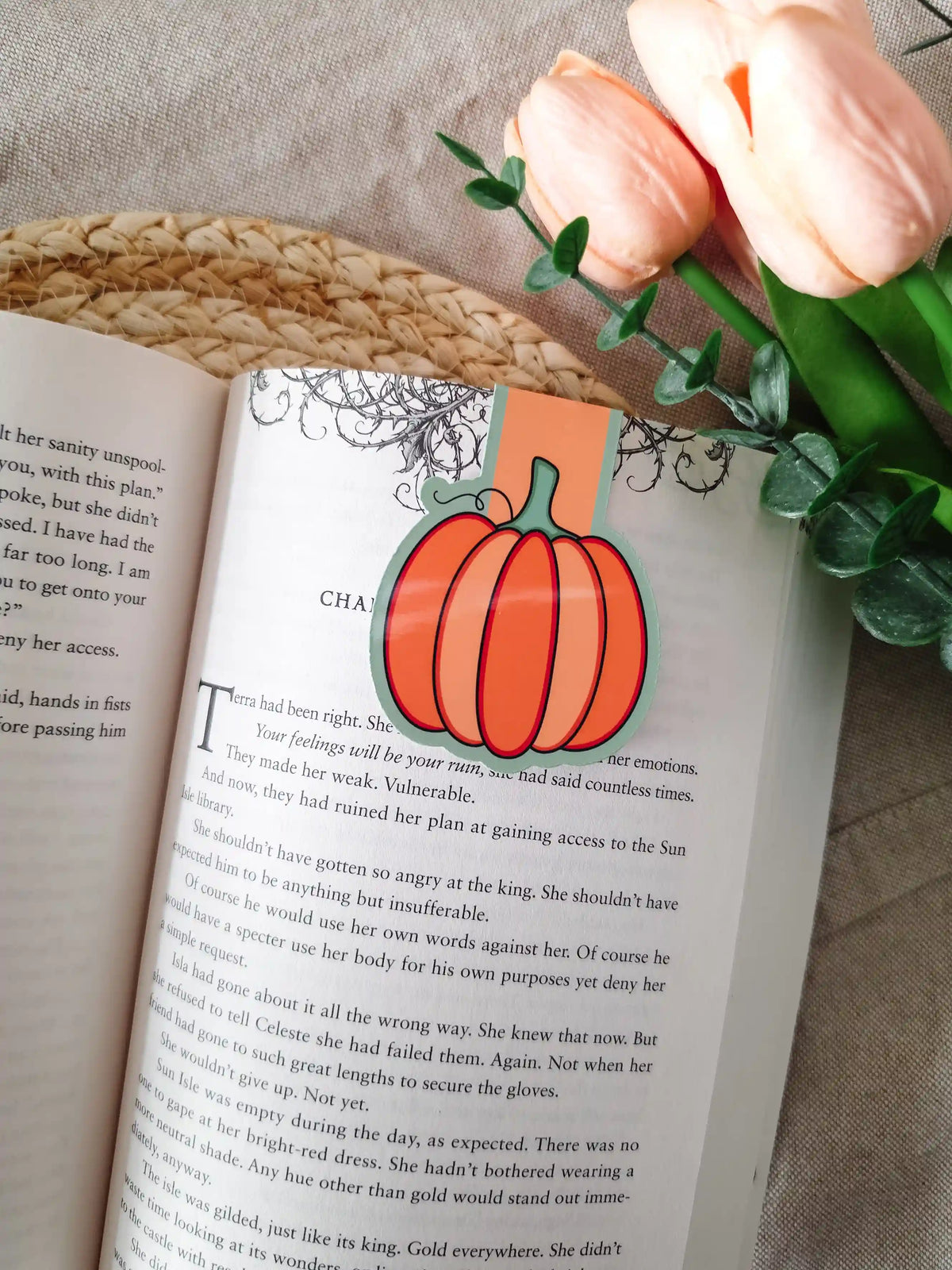 A die-cut magnetic bookmark in the shape of a round orange pumpkin with a green stem and dark outline, clipped onto the top corner of an open book resting on a woven rattan mat, with peach tulips and green eucalyptus sprigs in the upper right corner.