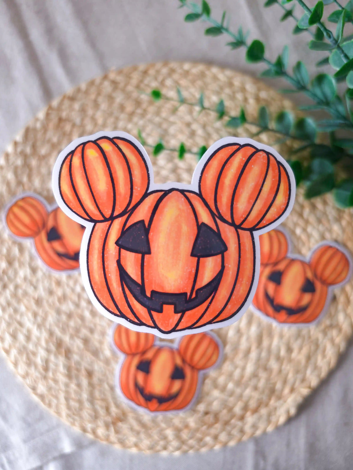 A large die-cut glossy vinyl sticker of a carved jack-o'-lantern pumpkin shaped like iconic round mouse ears, with a grinning carved face, displayed alongside two smaller versions of the same sticker on a woven rattan mat, with green eucalyptus sprigs in the upper right corner.