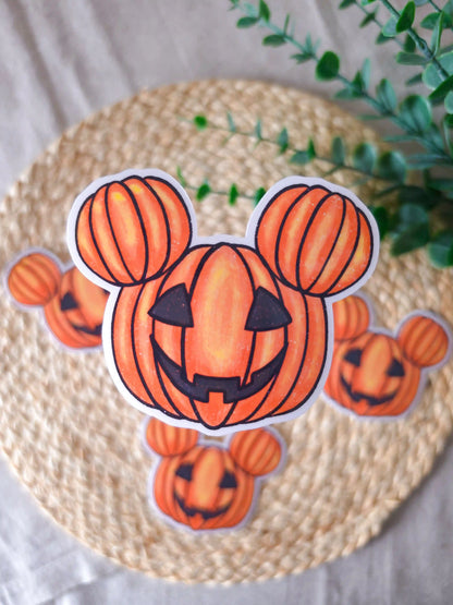 A large die-cut glossy vinyl sticker of a carved jack-o'-lantern pumpkin shaped like iconic round mouse ears, with a grinning carved face, displayed alongside two smaller versions of the same sticker on a woven rattan mat, with green eucalyptus sprigs in the upper right corner.