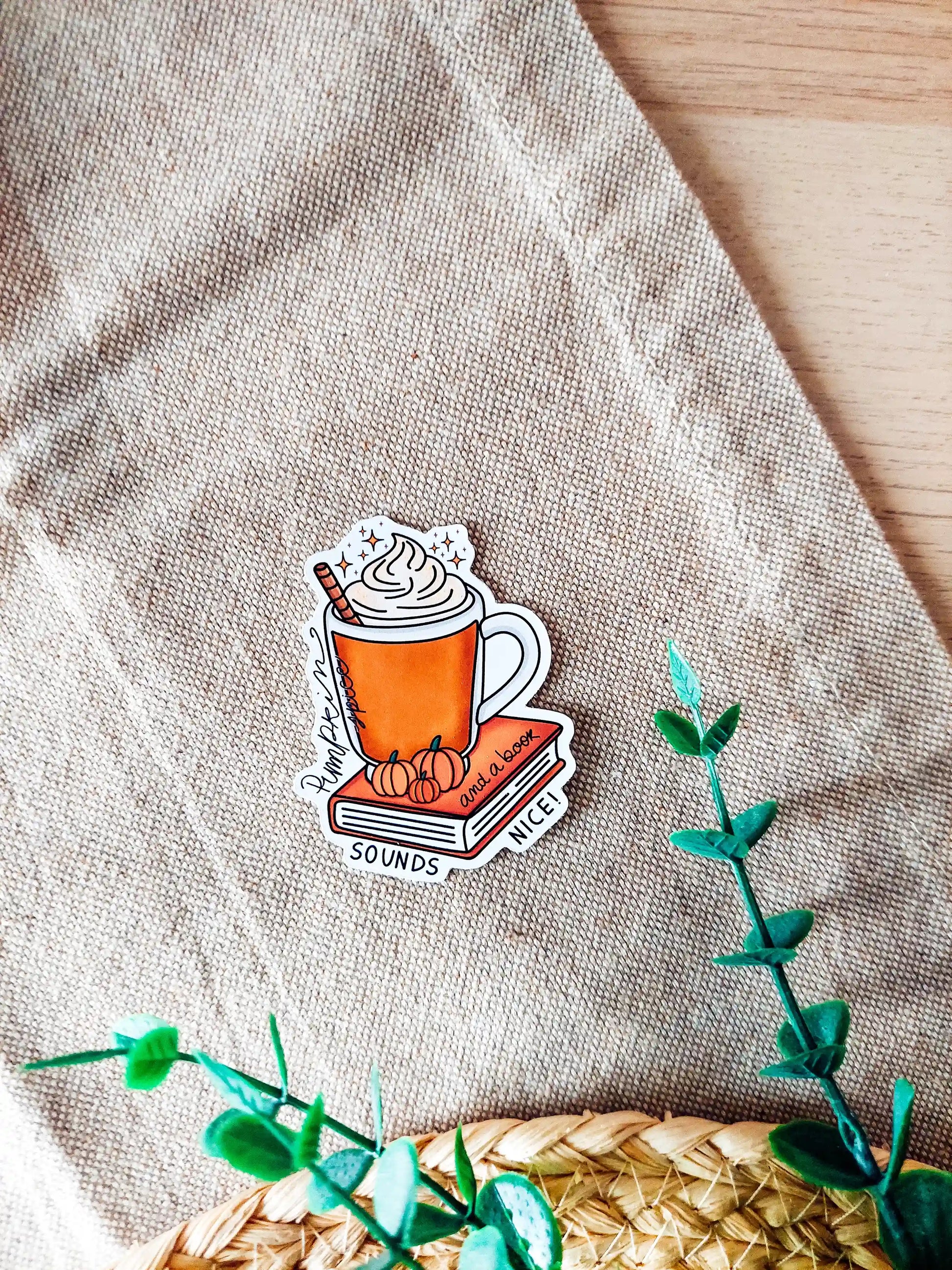 Vinyl sticker featuring an orange mug with whipped cream on top of a red book with small pumpkins and the tex "Pumpkin spice and a book sounds nice", photographed on neutral fabric background with woven mat.