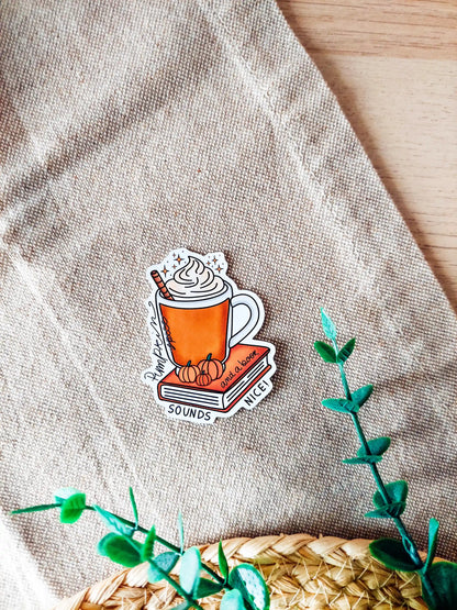 Vinyl sticker featuring an orange mug with whipped cream on top of a red book with small pumpkins and the tex "Pumpkin spice and a book sounds nice", photographed on neutral fabric background with woven mat.