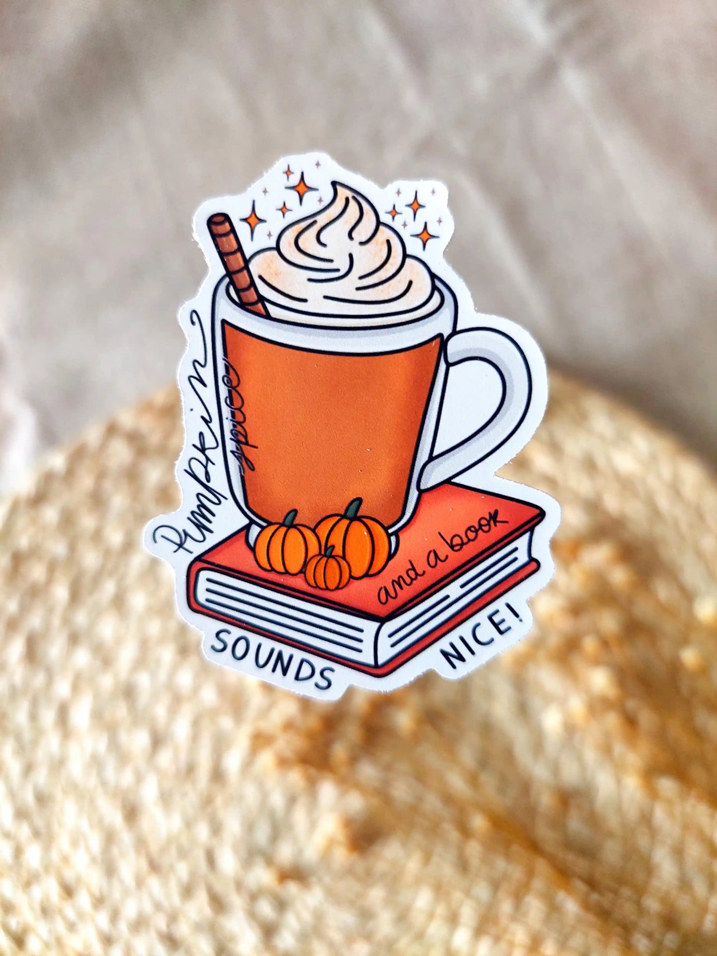 Vinyl sticker featuring an orange mug with whipped cream on top of a red book with small pumpkins and the tex "Pumpkin spice and a book sounds nice", photographed on neutral fabric background with woven mat.