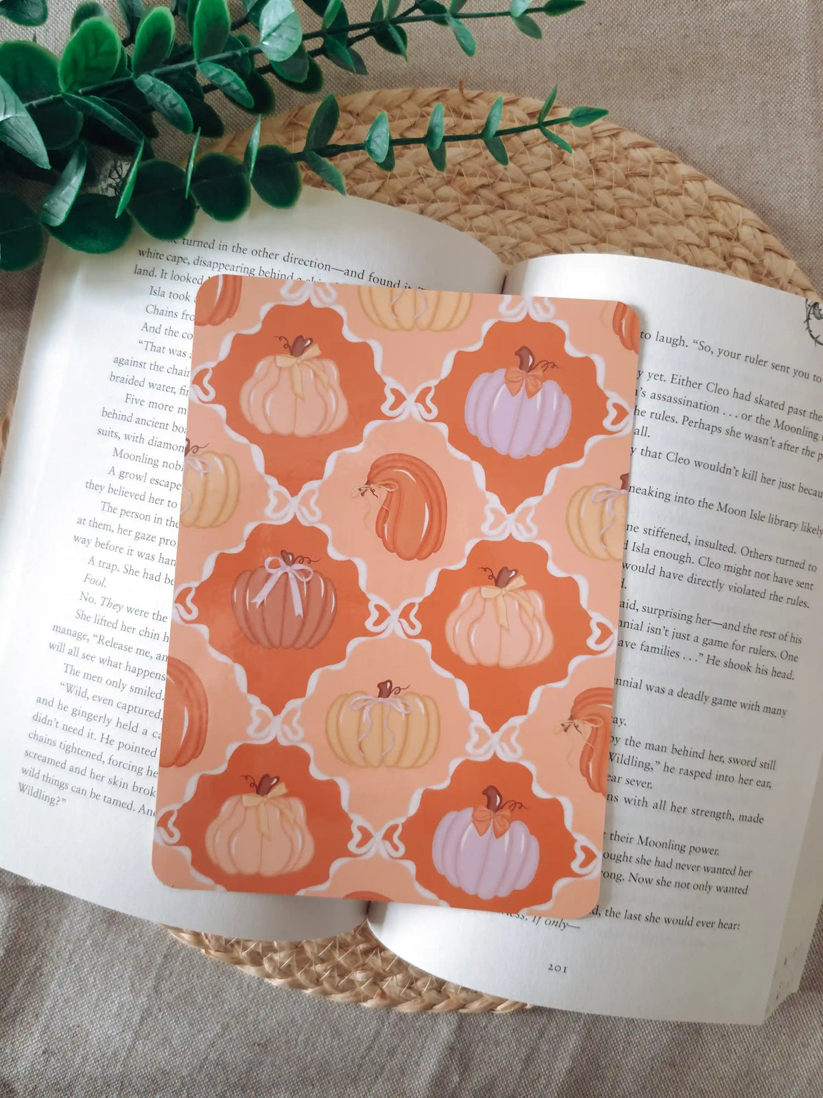 A laminated e-reader insert featuring a warm orange and terracotta tile pattern with illustrated pumpkins in peach, lavender, and blush tones, some decorated with small bows, resting on an open book on a woven rattan mat, with green eucalyptus sprigs in the upper right corner.