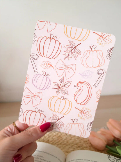 A laminated e-reader insert on a pale blush pink background featuring line-art illustrations of pumpkins in various shapes, autumn maple leaves, and decorative bows in warm orange, terracotta, and brown gradient tones, resting on an open book on a woven rattan mat, with green eucalyptus sprigs in the upper right corner.