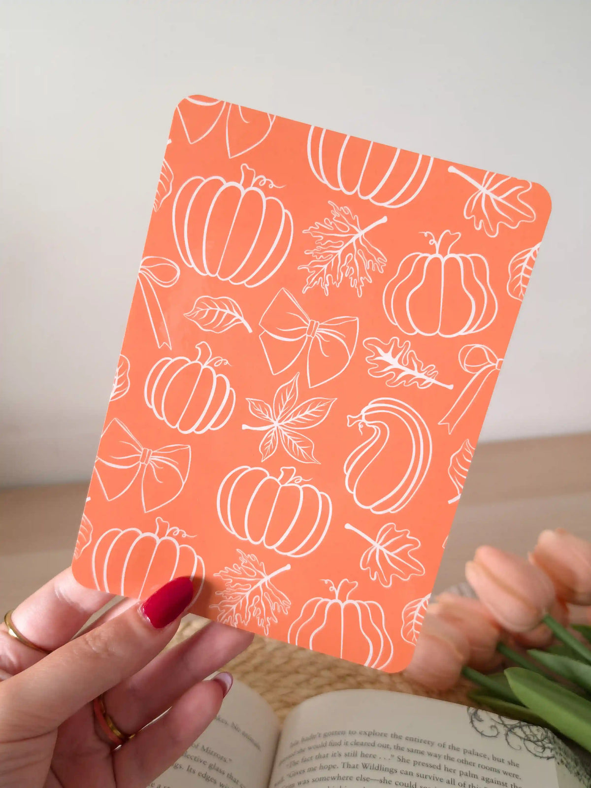 A laminated e-reader insert with a solid terracotta orange background featuring white line-art illustrations of pumpkins in various shapes, autumn maple leaves, and decorative bows scattered across the design, resting on an open book on a woven rattan mat, with green eucalyptus sprigs in the upper right corner.