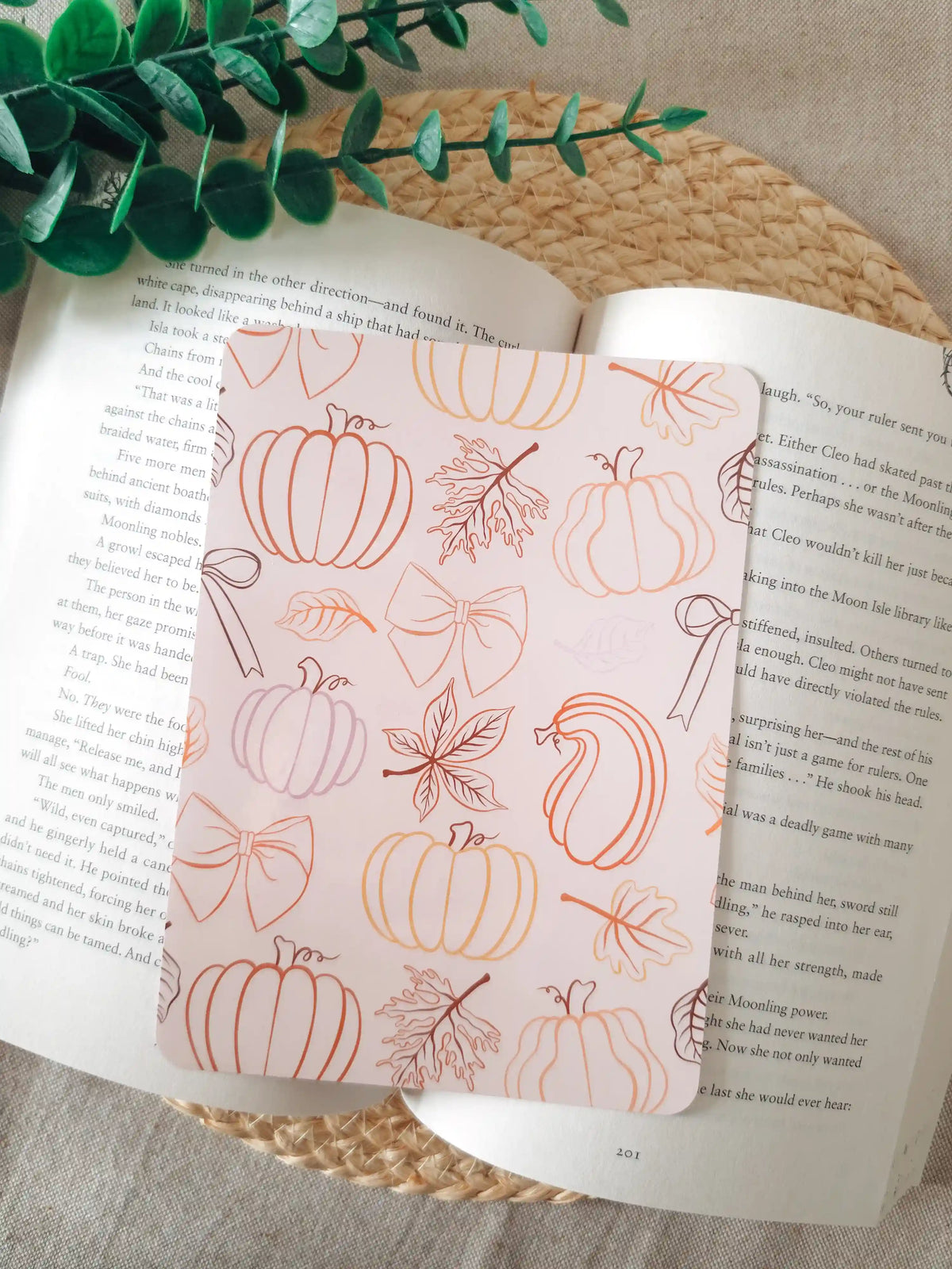 A laminated e-reader insert on a pale blush pink background featuring line-art illustrations of pumpkins in various shapes, autumn maple leaves, and decorative bows in warm orange, terracotta, and brown gradient tones, resting on an open book on a woven rattan mat, with green eucalyptus sprigs in the upper right corner.