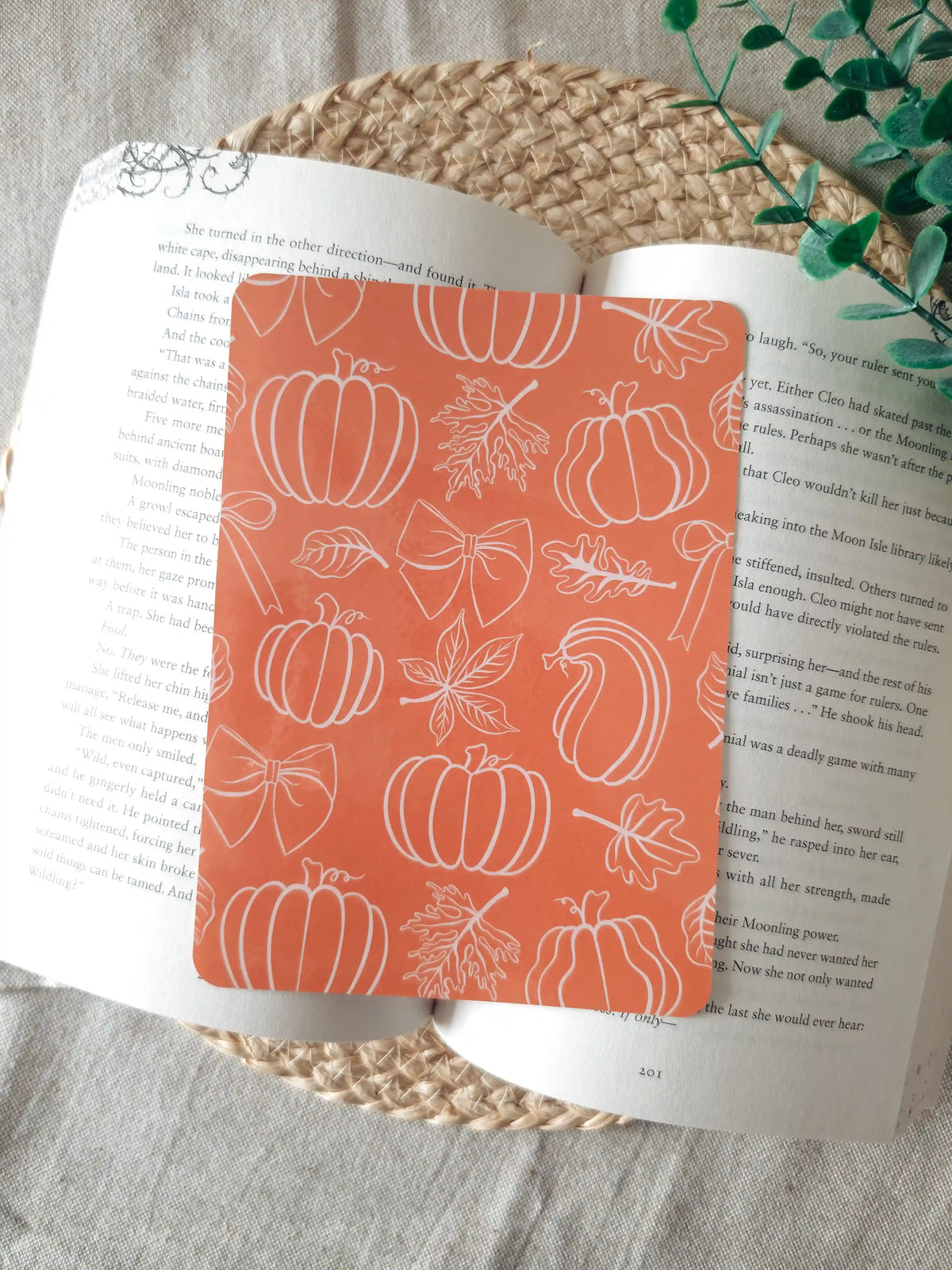 A laminated e-reader insert with a solid terracotta orange background featuring white line-art illustrations of pumpkins in various shapes, autumn maple leaves, and decorative bows scattered across the design, resting on an open book on a woven rattan mat, with green eucalyptus sprigs in the upper right corner.