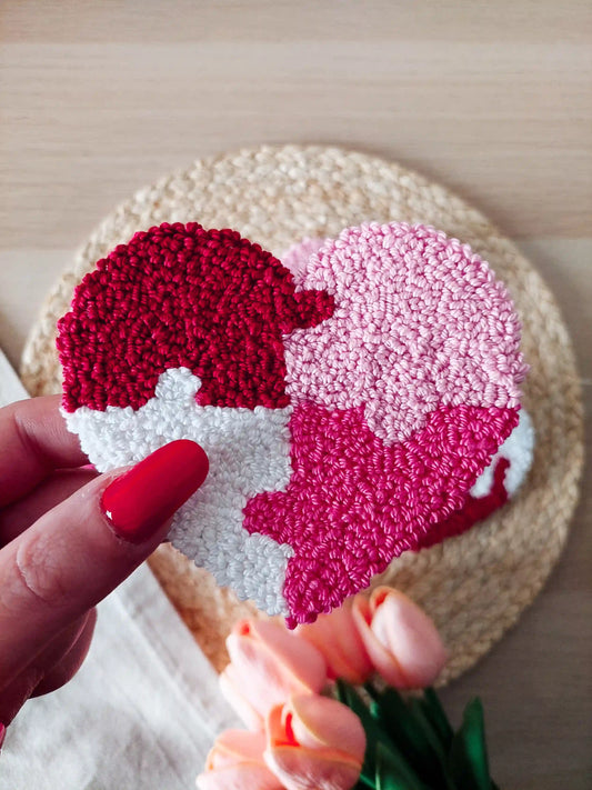 Handmade punch needle puzzle heart coaster in red, pink and white photographed on a woven mat with decorative flowers in the back.