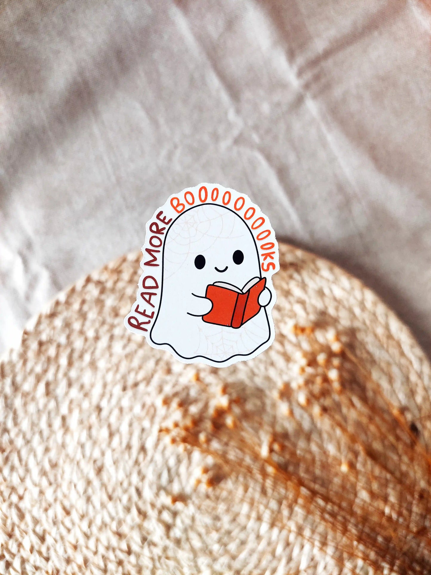 Vinyl sticker of a cute ghost reading a book with the phrase "Read More Booooooks" in lettering, photographed on a neutral fabric background.