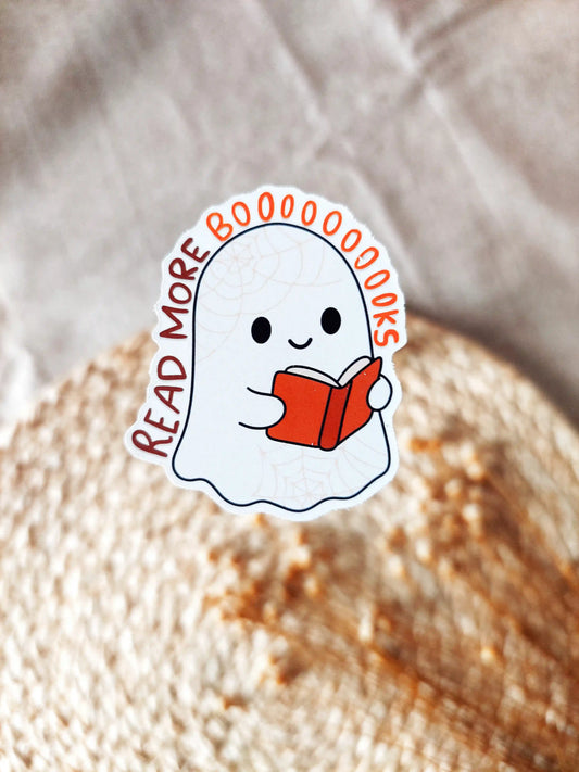 Vinyl sticker of a cute ghost reading a book with the phrase "Read More Booooooks" in lettering, photographed on a neutral fabric background.