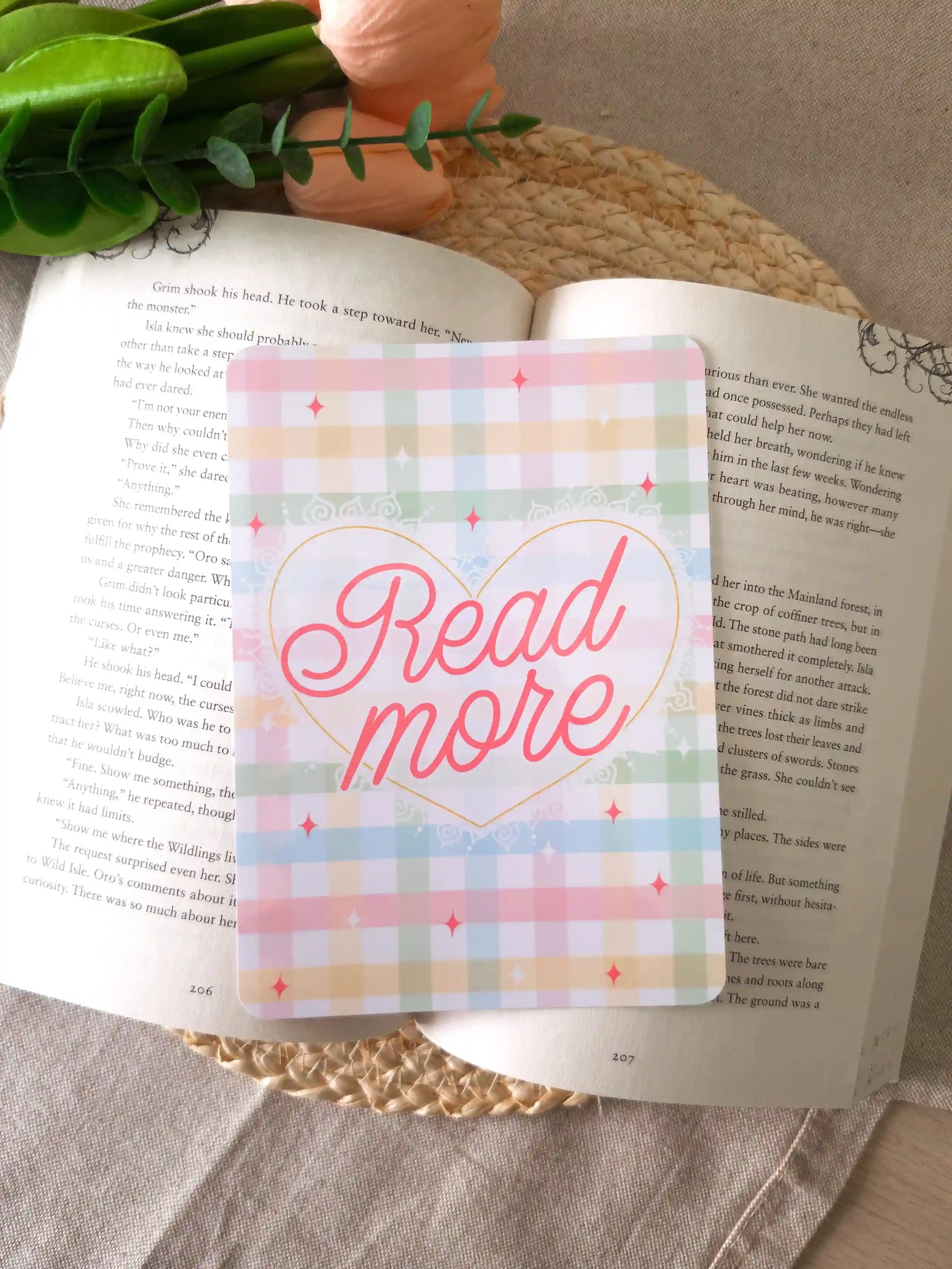 An e-reader insert with a pastel gingham background in soft pink, blue, green, and yellow, featuring a gold heart outline with the text "Read More" in pink cursive script and pink sparkle details, placed on an open book resting on a woven rattan mat.