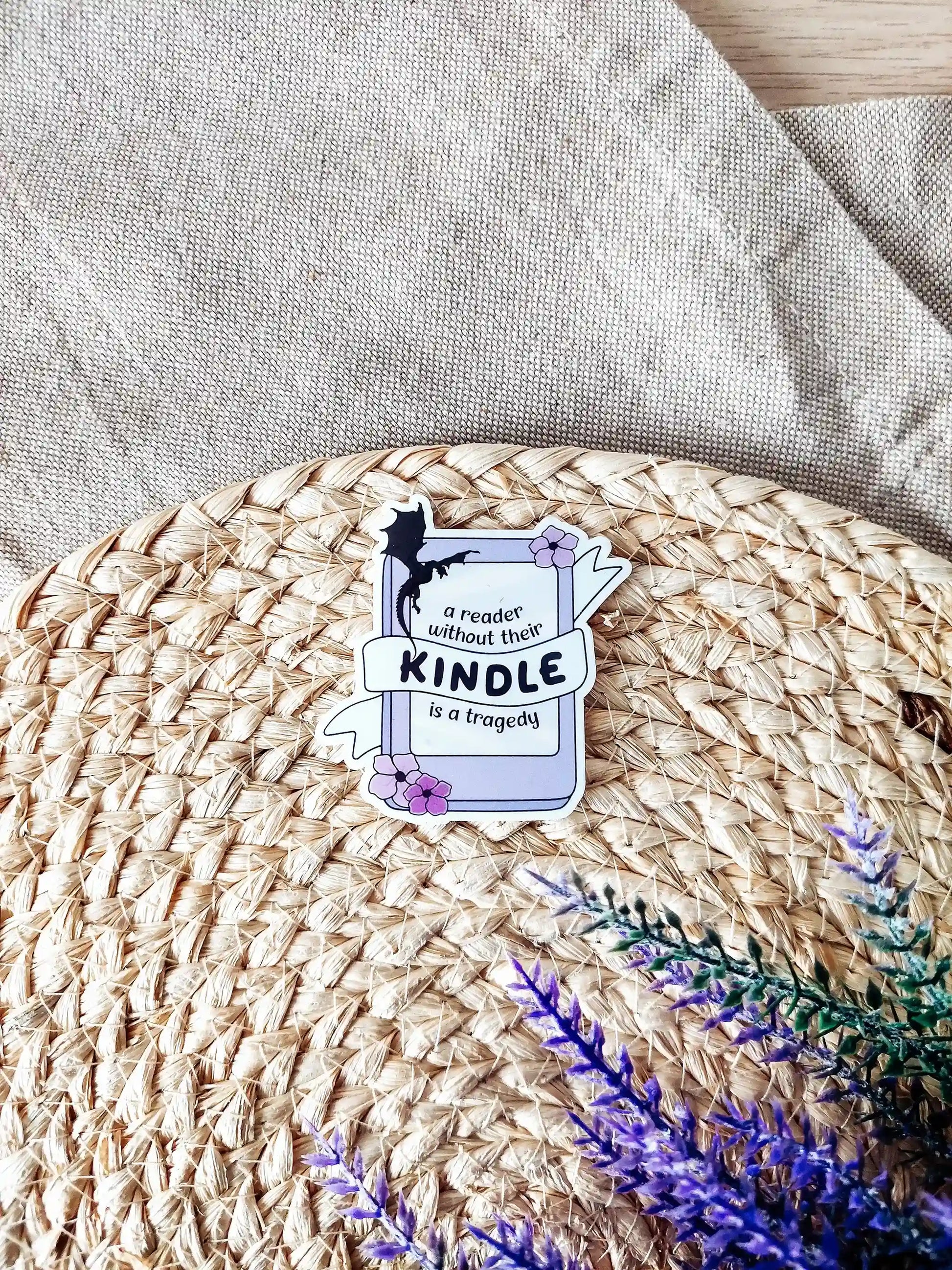 Vinyl sticker of a kinddle with flowers and a dragon silhouette, featuring the phrase "A reader without their Kindle is a tragedy", photographed on a woven neutral background.