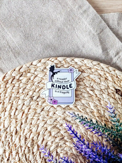 Vinyl sticker of a kinddle with flowers and a dragon silhouette, featuring the phrase "A reader without their Kindle is a tragedy", photographed on a woven neutral background.