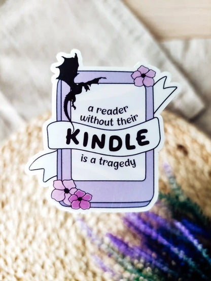 Vinyl sticker of a kinddle with flowers and a dragon silhouette, featuring the phrase "A reader without their Kindle is a tragedy", photographed on a woven neutral background.