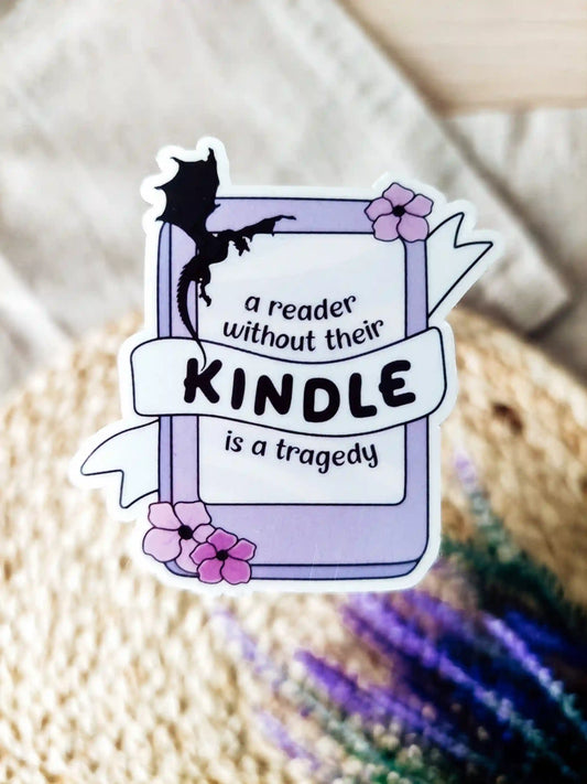 Vinyl sticker of a kinddle with flowers and a dragon silhouette, featuring the phrase "A reader without their Kindle is a tragedy", photographed on a woven neutral background.