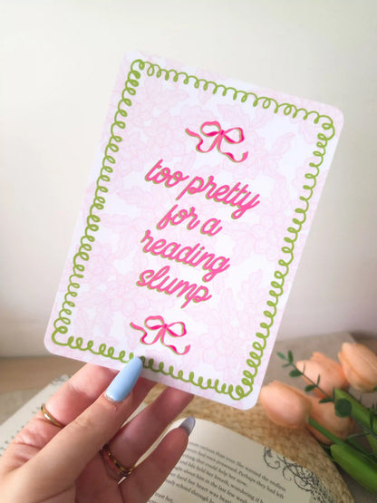 A hand holding an e-reader insert with a pink lacey background, green wavy decorative border, two pink bow details, and the text "too pretty for a reading slump" in bold pink cursive script.