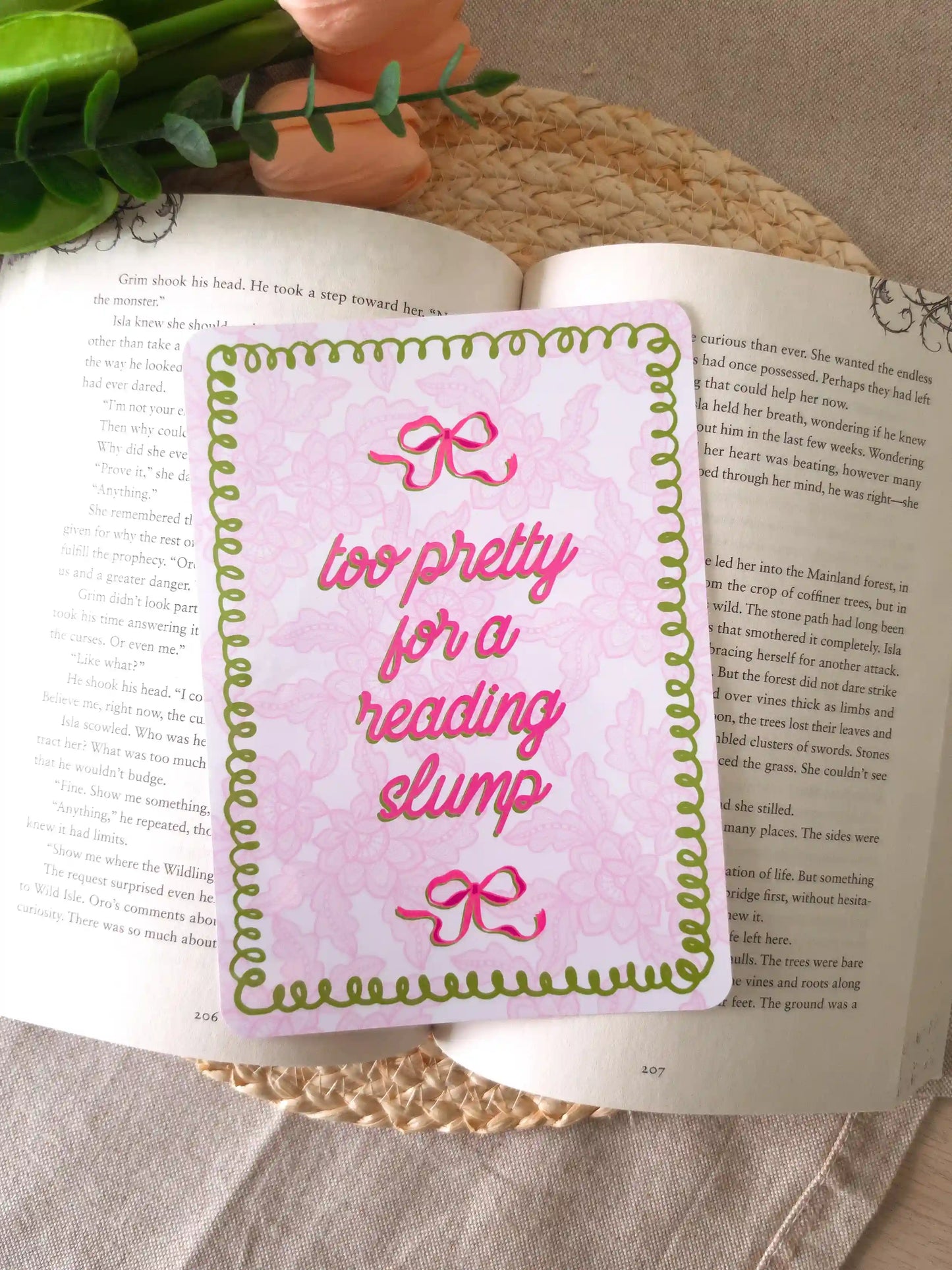 An e-reader insert with a pink lacey background, green wavy decorative border, two pink bow details, and the text "too pretty for a reading slump" in bold pink cursive script, placed on an open book resting on a woven rattan mat. 