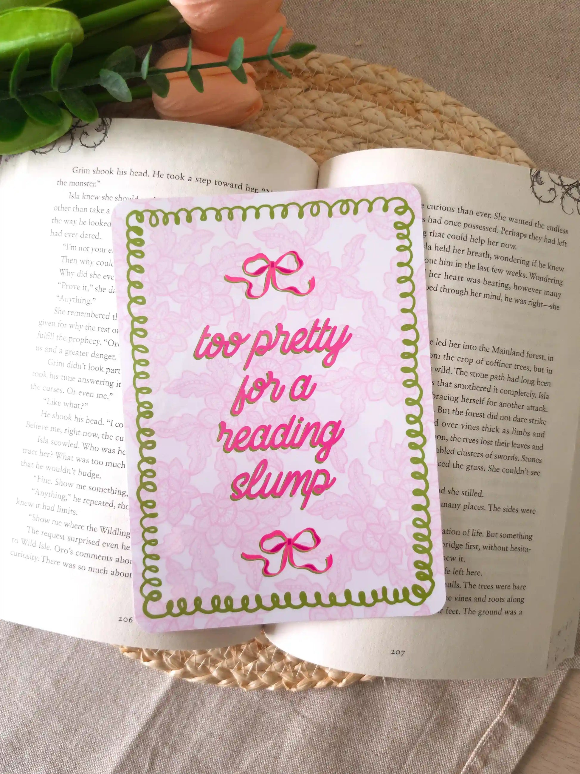 An e-reader insert with a pink lacey background, green wavy decorative border, two pink bow details, and the text "too pretty for a reading slump" in bold pink cursive script, placed on an open book resting on a woven rattan mat. 