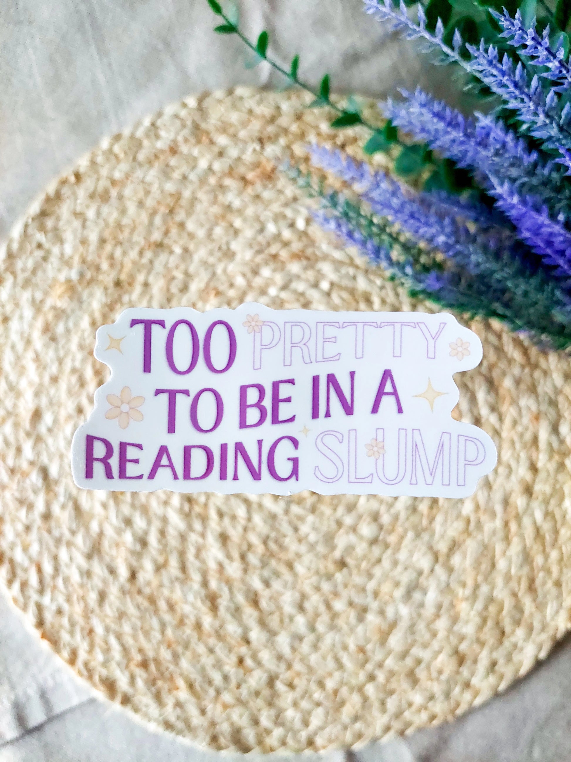 "Too Pretty To Be In A Reading Slump" sticker featuring purple and lilac text with small flowers and stars details on a woven background.