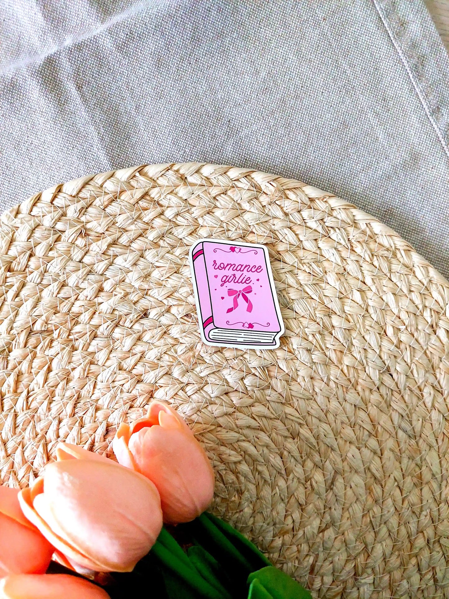 Vinyl sticker featuring a pink book with bow and heart details on the cover and the text "Romance girlie", photographed on neutral fabric background with woven mat.