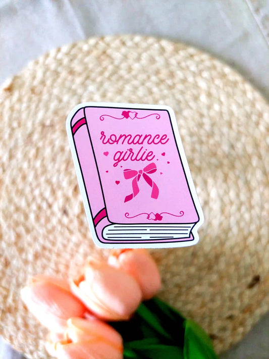 Vinyl sticker featuring a pink book with bow and heart details on the cover and the text "Romance girlie", photographed on neutral fabric background with woven mat.