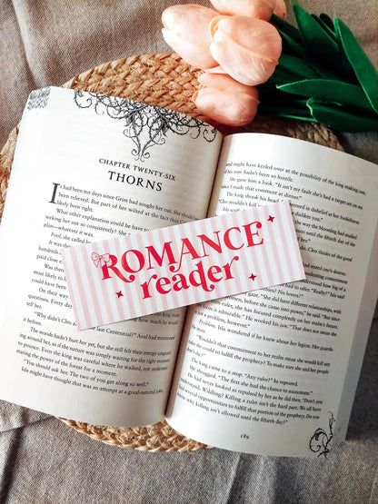 Illustrated bookmark with the text "Romance reader", featuring pink stripes and decorative stars, resting on an open book.
