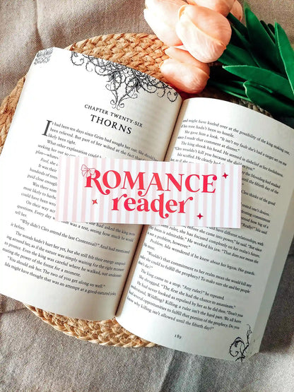 Illustrated bookmark with the text "Romance reader", featuring pink stripes and decorative stars, resting on an open book.